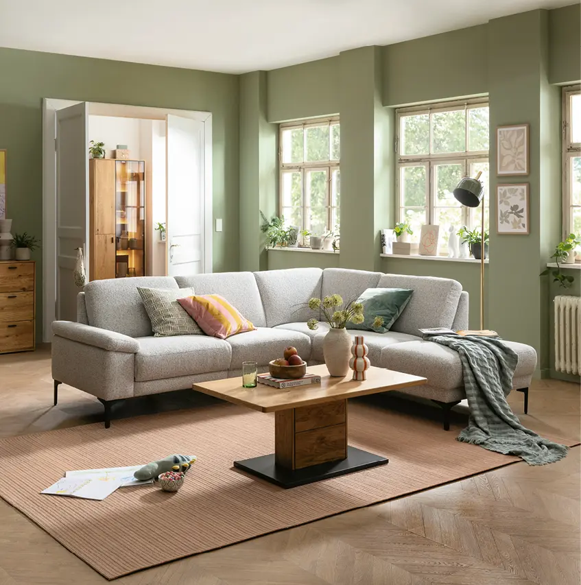 Helles Wohnzimmer mit großem Ecksofa in Stoffoptik, Holzcouchtisch und grüner Wandfarbe. Natürliche Einrichtung mit Wohnaccessoires, Pflanzen und viel Tageslicht für ein modernes und gemütliches Wohnambiente.